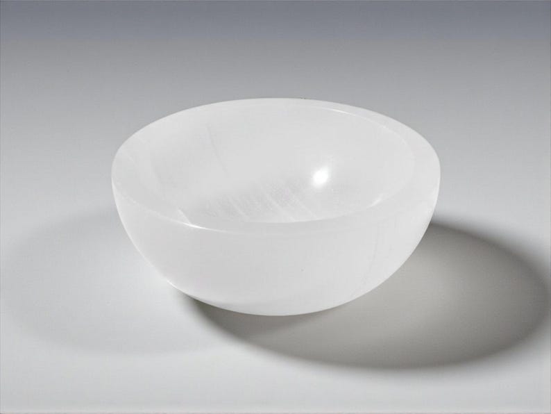 May include: A white, oval bowl with a smooth, polished surface. The bowl is made of a translucent material, and it casts a shadow on the surface below. The bowl is likely used for decorative purposes.