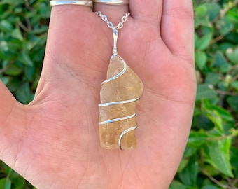 May include: A silver chain necklace with a raw, light-yellow citrine crystal pendant. The crystal is wrapped in silver wire. The necklace is displayed on an open hand, with green foliage in the background.