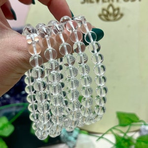 May include: Clear quartz crystal bead bracelets. Each bracelet is composed of multiple strands of clear, round beads. The beads are strung together, creating a layered effect. The bracelets are held in a hand with green nail polish. The background is blurred.