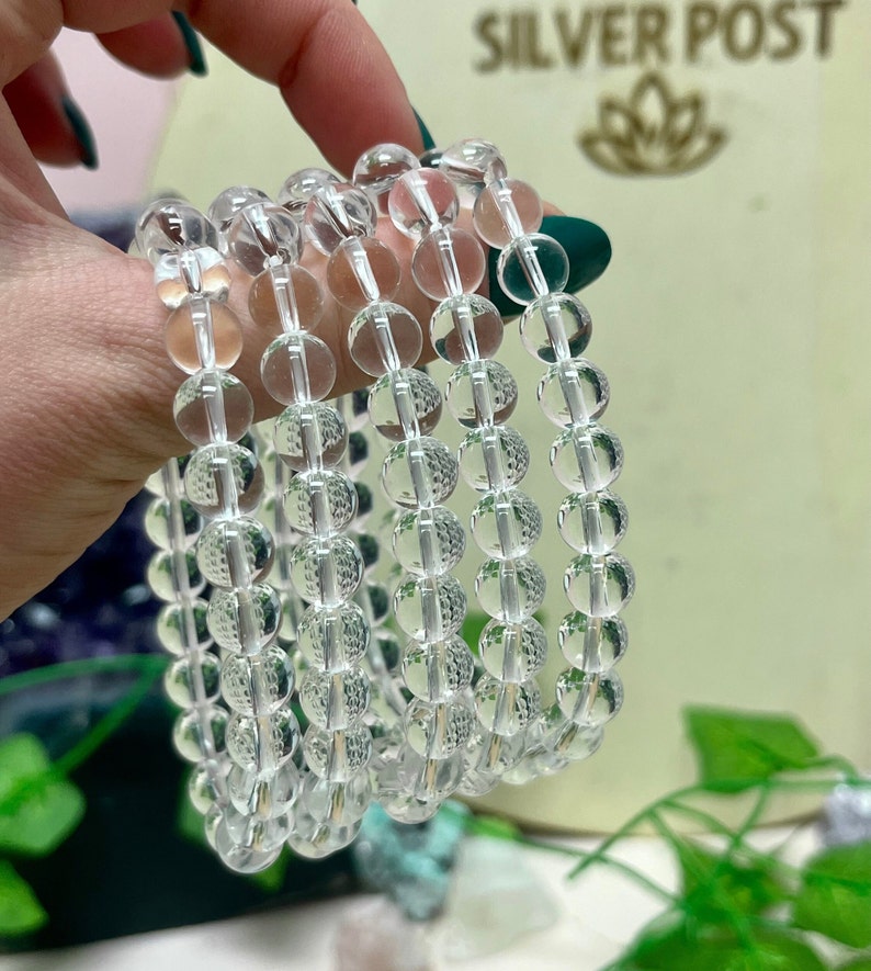 May include: Clear quartz crystal bead bracelets. Each bracelet is composed of multiple strands of clear, round beads. The beads are strung together, creating a layered effect. The bracelets are held in a hand with green nail polish. The background is blurred.