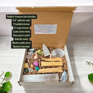 May include: An open cardboard box filled with a variety of crystals, stones, and other items. The box includes a selenite ruler, tumbled stones, rough stones, palo santo sticks, a rose quartz point, a selenite tower, a palm stone, and a keychain. The box is labelled "Radiant Treasure Box (Medium)" and lists the contents of the box.