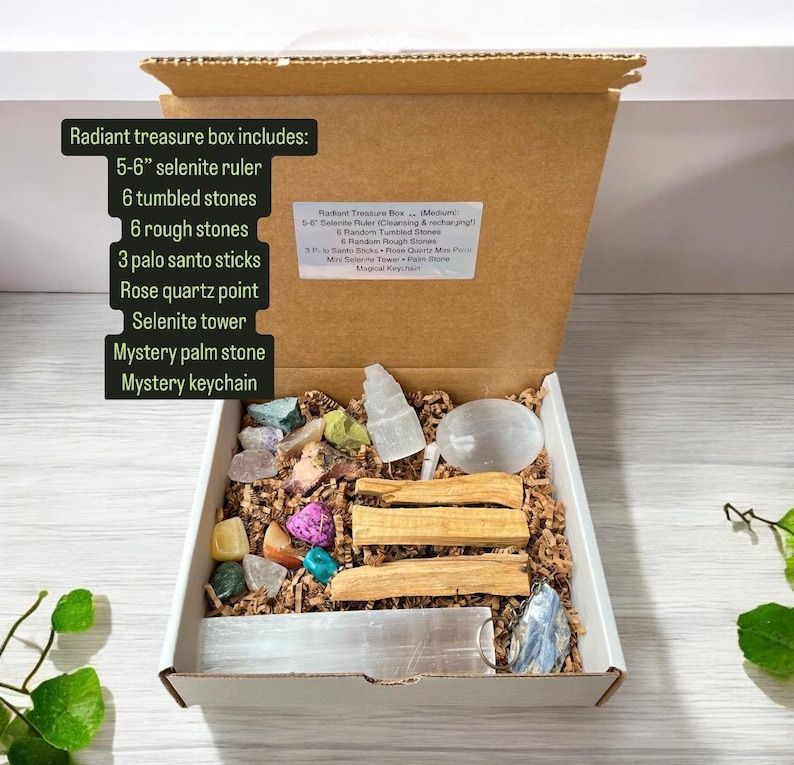May include: An open cardboard box filled with a variety of crystals, stones, and other items. The box includes a selenite ruler, tumbled stones, rough stones, palo santo sticks, a rose quartz point, a selenite tower, a palm stone, and a keychain. The box is labelled "Radiant Treasure Box (Medium)" and lists the contents of the box.