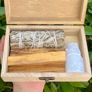 May include: A wooden box with a hinged lid, open to reveal a set of smudging tools. The box contains white sage bundles, palo santo sticks, and a selenite crystal.