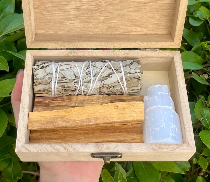 May include: A wooden box with a hinged lid, open to reveal a set of smudging tools. The box contains white sage bundles, palo santo sticks, and a selenite crystal.