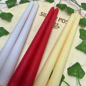 May include: Three pairs of taper candles in white, red, and cream colors. The candles are arranged on a wooden surface with green leaves in the background.