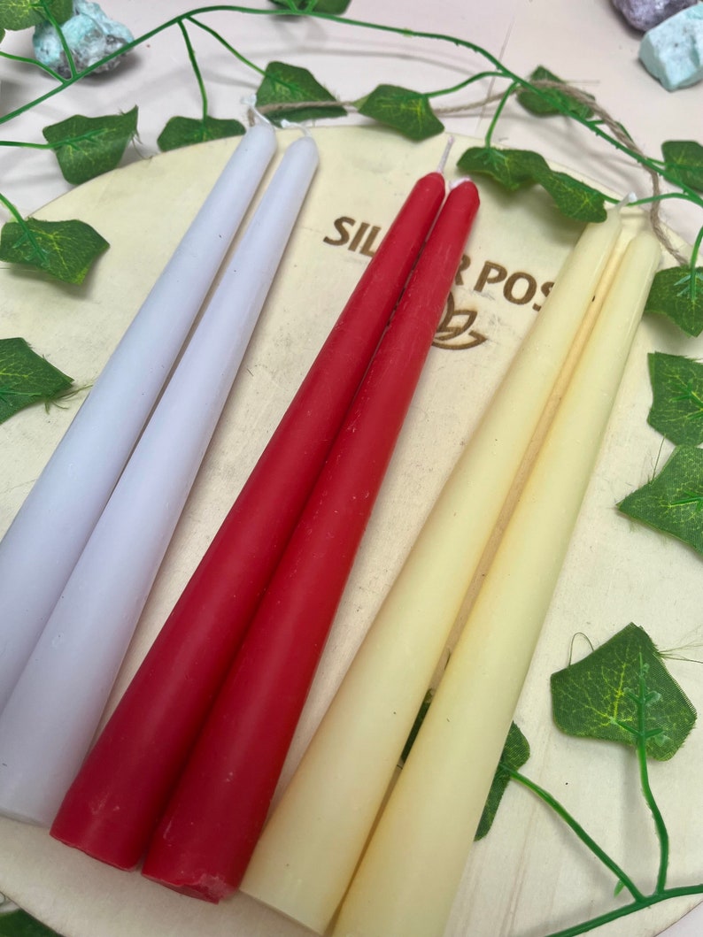 May include: Three pairs of taper candles in white, red, and cream colors. The candles are arranged on a wooden surface with green leaves in the background.