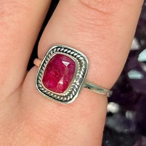 May include: A silver ring with a red gemstone set in a square bezel. The bezel has a rope-like design around the edge.