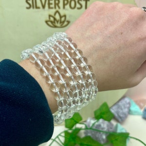 May include: Clear quartz crystal bead bracelets stacked on a wrist. The bracelets are made of translucent, round beads strung together. The background includes the text "SILVER POST" and a floral logo.