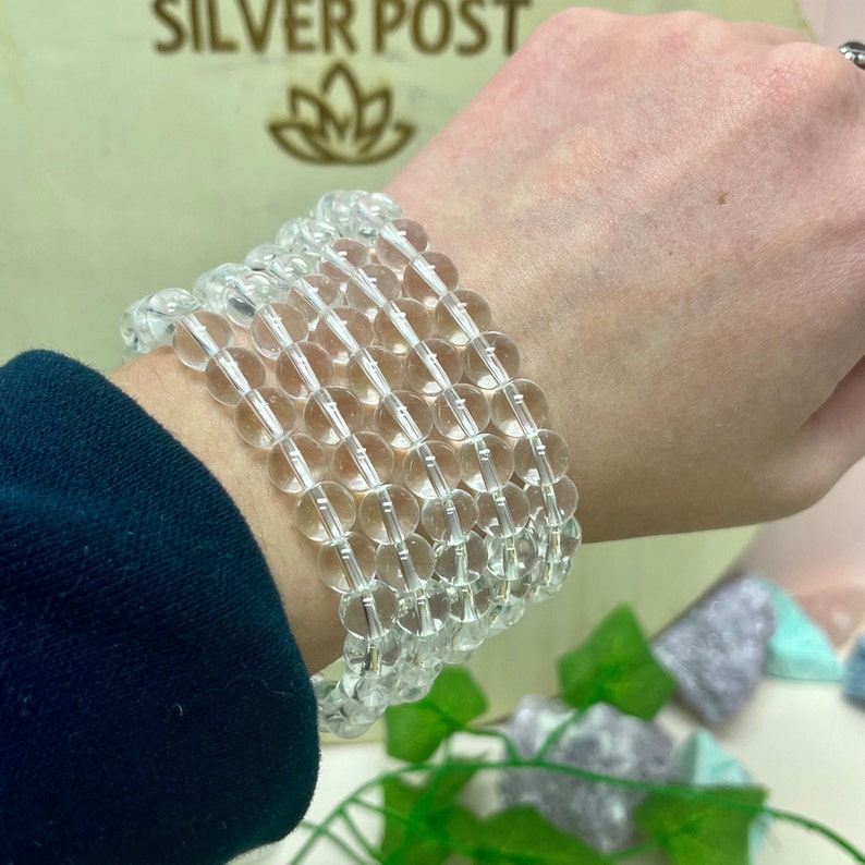 May include: Clear quartz crystal bead bracelets stacked on a wrist. The bracelets are made of translucent, round beads strung together. The background includes the text "SILVER POST" and a floral logo.