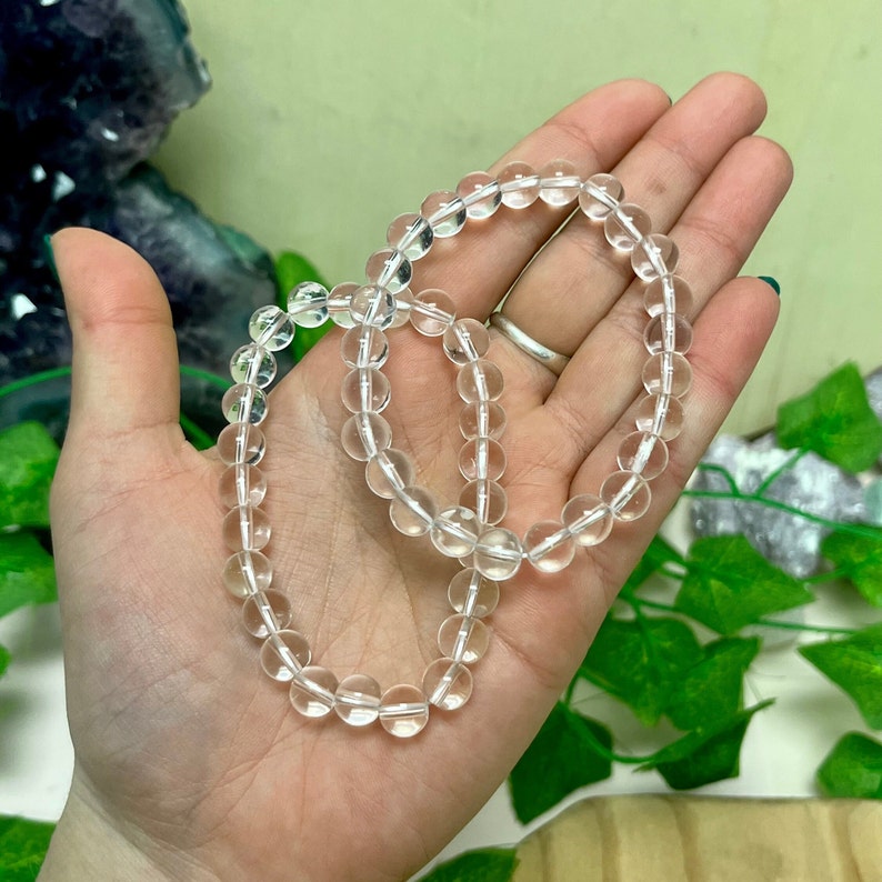 May include: Two clear quartz crystal bead bracelets. Each bracelet is composed of round, translucent beads strung together. The bracelets are held in an open hand, with a green and purple background.