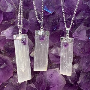 May include: Three silver necklaces with selenite crystal pendants and amethyst accents. The pendants are rectangular and have a smooth, white finish. The necklaces are displayed against a background of purple amethyst crystals.