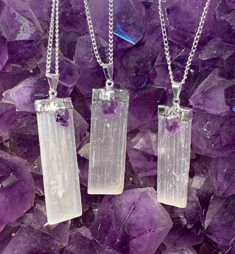 May include: Three silver necklaces with selenite crystal pendants and amethyst accents. The pendants are rectangular and have a smooth, white finish. The necklaces are displayed against a background of purple amethyst crystals.