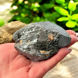 K&ouml;nnte beinhalten: Ein dunkelgraues, grob strukturiertes Mineralexemplar mit silbernen und braunen Flecken. Der Naturstein wird in einer Hand gehalten, mit gr&uuml;nem Laub im Hintergrund. Die Oberfl&auml;che des Gesteins hat einen metallischen Glanz.