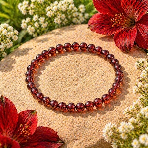 May include: A beaded bracelet made of small, round, deep red gemstones. The bracelet is resting on a sandy surface, surrounded by red flowers and small white flowers.