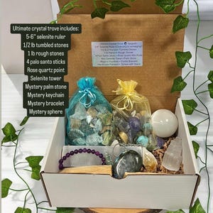 May include: A white box filled with various crystals and crystal tools. The box includes a selenite ruler, tumbled stones, rough stones, palo santo sticks, a rose quartz point, a selenite tower, a palm stone, a keychain, a bracelet, and a sphere. The box is labelled "Ultimate Crystal Trove".