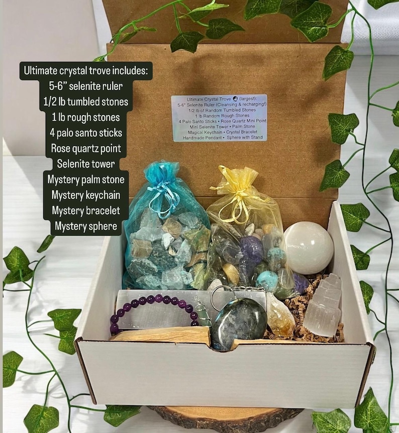 May include: A white box filled with various crystals and crystal tools. The box includes a selenite ruler, tumbled stones, rough stones, palo santo sticks, a rose quartz point, a selenite tower, a palm stone, a keychain, a bracelet, and a sphere. The box is labelled "Ultimate Crystal Trove".