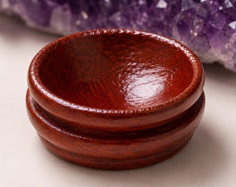 May include: A small, round, wooden bowl with a rich, reddish-brown finish. The bowl has a decorative, ridged edge and sits on a slightly wider base. The wood grain is visible, adding texture. A purple crystal formation is in the background.
