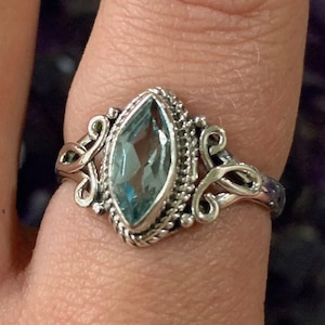 May include: A silver ring with a blue gemstone set in a marquise shape. The ring has a delicate, ornate design with swirls and curves.