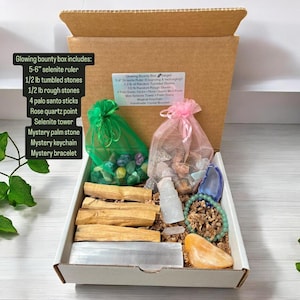 May include: A white box filled with crystals, stones, and palo santo sticks. The box includes a selenite ruler, tumbled stones, rough stones, a rose quartz point, a selenite tower, a palm stone, a keychain, and a bracelet. The box is labelled "Glowing Bounty Box".