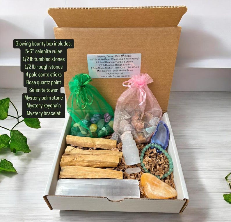 May include: A white box filled with crystals, stones, and palo santo sticks. The box includes a selenite ruler, tumbled stones, rough stones, a rose quartz point, a selenite tower, a palm stone, a keychain, and a bracelet. The box is labelled "Glowing Bounty Box".