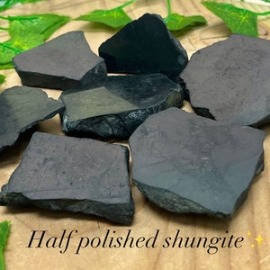 May include: A collection of seven half-polished black shungite stones of various sizes and shapes. The stones have a rough, natural texture on one side and a smooth, polished surface on the other.