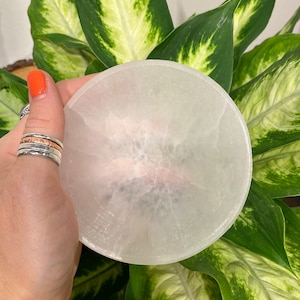 May include: A circular, white selenite charging plate. The plate is held in a hand with orange nail varnish and silver and rose gold rings. Green and white patterned leaves are in the background.