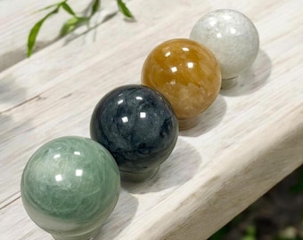 May include: Four decorative stone spheres in a row. The spheres are green, dark teal, amber, and white. Each sphere has a rounded top and a short, cylindrical base. The spheres are arranged on a light-colored wooden surface.
