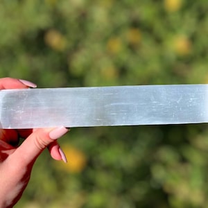 May include: A smooth, white selenite wand with a rectangular shape. The wand is held in a hand against a blurred green background.