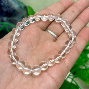 May include: Clear quartz crystal bead bracelet. The bracelet is made of round, transparent quartz beads strung together on a white elastic cord. The beads are approximately 0.39 inches in diameter. The bracelet is shown on a person's hand.