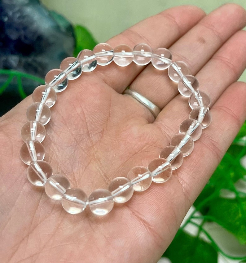 May include: Clear quartz crystal bead bracelet. The bracelet is made of round, transparent quartz beads strung together on a white elastic cord. The beads are approximately 0.39 inches in diameter. The bracelet is shown on a person's hand.