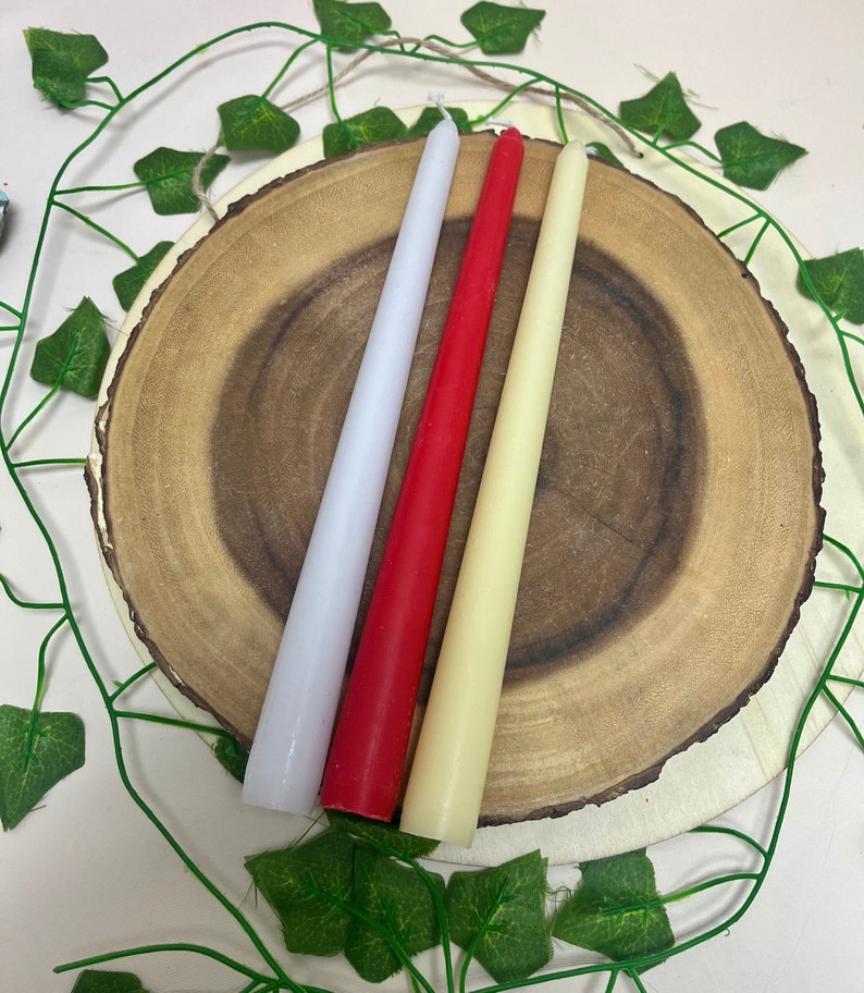May include: Three taper candles on a wooden slice. The candles are white, red, and cream.