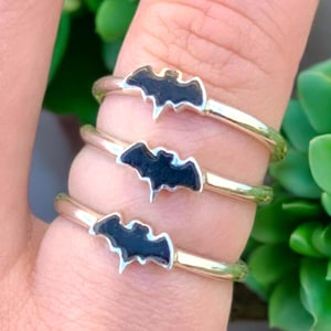 May include: Three silver rings with black bat charms. The rings are stacked on a finger.