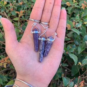 May include: Three purple amethyst crystal point pendants with silver chain and bail. The pendants are held in a hand.