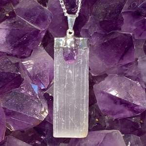 May include: A silver chain necklace with a white selenite crystal pendant and a small amethyst crystal at the top. The pendant is set in a silver bezel and hangs from a silver chain.