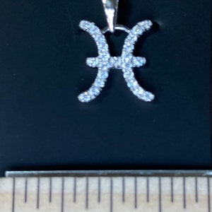 K&ouml;nnte beinhalten: Silberkette mit einem Anh&auml;nger in Form des Sternzeichen Fische. Der Anh&auml;nger ist aus Silber und hat kleine, klare Steine, die in Form des Fische-Symbols gesetzt sind. Die Kette ist etwa 2,5 cm lang.