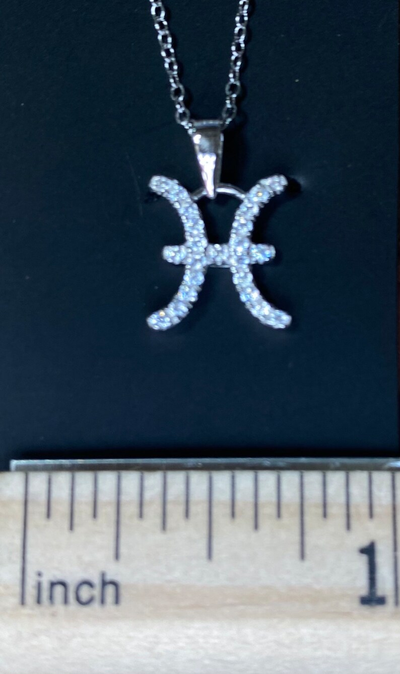K&ouml;nnte beinhalten: Silberkette mit einem Anh&auml;nger in Form des Sternzeichen Fische. Der Anh&auml;nger ist aus Silber und hat kleine, klare Steine, die in Form des Fische-Symbols gesetzt sind. Die Kette ist etwa 2,5 cm lang.