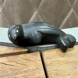 May include: A black carved stone sculpture of a seal. The seal is lying on its belly with its head turned to the side. The sculpture has white eyes.