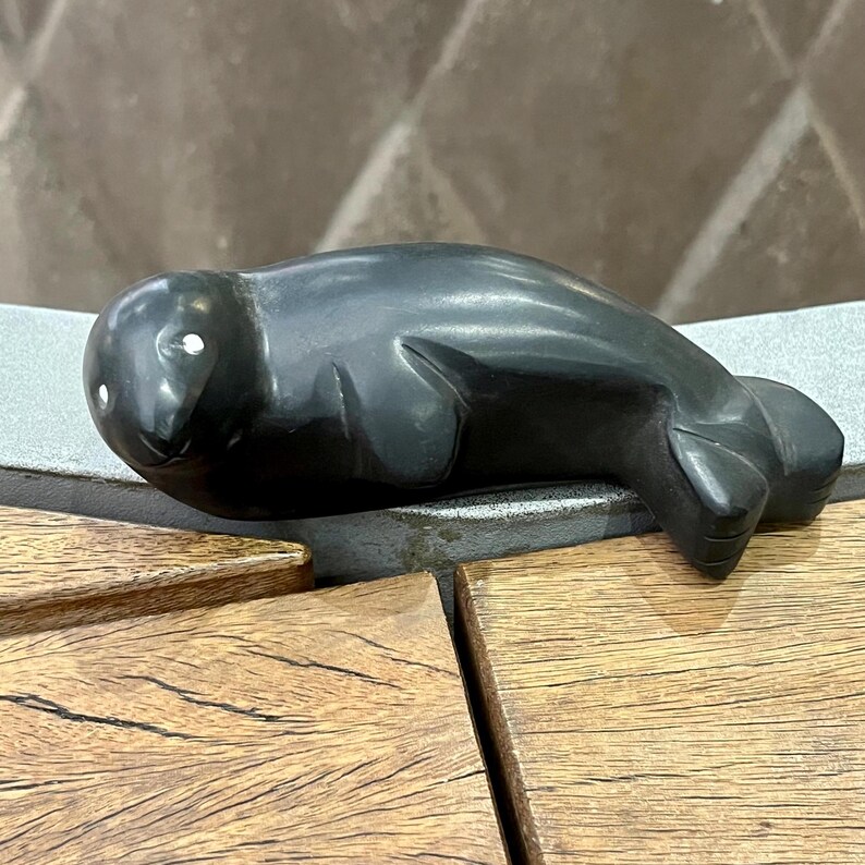 May include: A black carved stone sculpture of a seal. The seal is lying on its belly with its head turned to the side. The sculpture has white eyes.