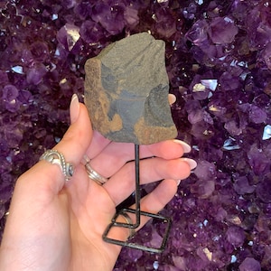 May include: A dark gray and brown stone with a rough texture is displayed on a black metal stand. The stand has a square base and a single upright post.