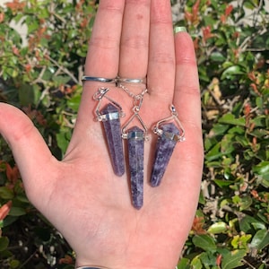 May include: Three purple amethyst crystal point pendants on silver chains. The pendants are in the shape of a long, thin point with a silver cap at the top. The chains are attached to the silver caps.