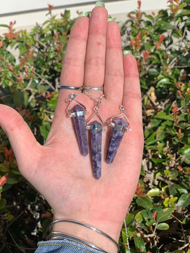 May include: Three purple amethyst crystal point pendants on silver chains. The pendants are in the shape of a long, thin point with a silver cap at the top. The chains are attached to the silver caps.