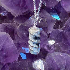 May include: A silver chain necklace with a black and white crystal pendant wrapped in silver wire. The pendant is displayed on a bed of purple amethyst crystals.