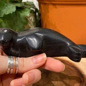 May include: A black, hand-carved stone sculpture of a seal. The sculpture is smooth and polished, with a realistic depiction of the animal.