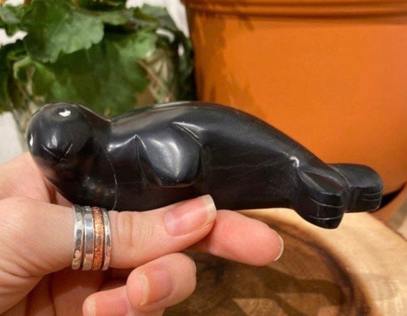 May include: A black, hand-carved stone sculpture of a seal. The sculpture is smooth and polished, with a realistic depiction of the animal.