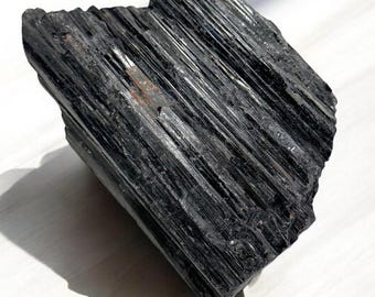 May include: A raw, black tourmaline crystal with a striated texture. The natural gemstone has a glossy sheen and is displayed on a white surface. The crystal's dark color and unique form make it a striking mineral specimen.
