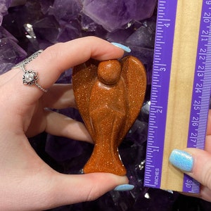 May include: A small, golden brown, carved angel figurine made of sunstone. The angel is approximately 5 cm tall and is being held in a hand for size reference.