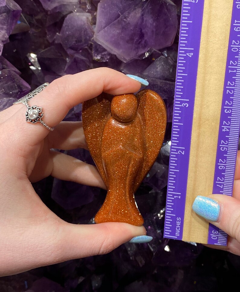 May include: A small, golden brown, carved angel figurine made of sunstone. The angel is approximately 5 cm tall and is being held in a hand for size reference.