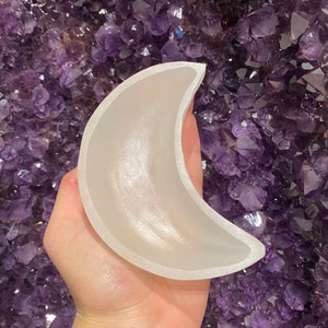 May include: A crescent moon-shaped object made of a translucent, frosted material. The moon has a smooth, curved surface and a defined edge. The background is a cluster of deep purple crystals.