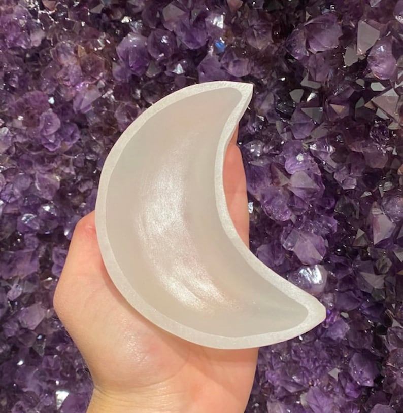 May include: A crescent moon-shaped object made of a translucent, frosted material. The moon has a smooth, curved surface and a defined edge. The background is a cluster of deep purple crystals.