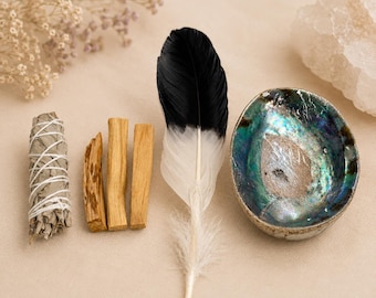 May include: An assortment of spiritual items, including a white sage bundle, palo santo sticks, a black and white feather, and an abalone shell. The items are arranged on a neutral-toned surface, suggesting a focus on natural elements and rituals.
